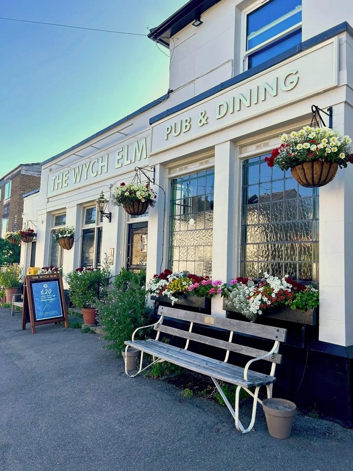 The white facade of The Wych Elm pub and dining room, with hanging flower baskets and a bench outside